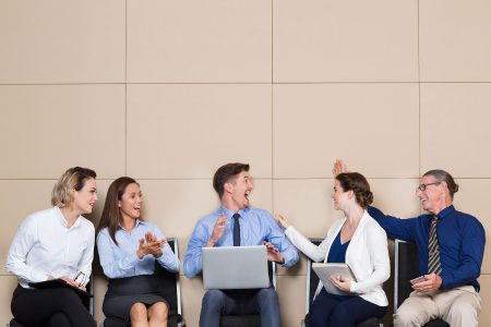 Five cheerful business people sitting in row at wall in waiting room. Young businessman with laptop is cheering ecstatically. Others are congratulating him and smiling.